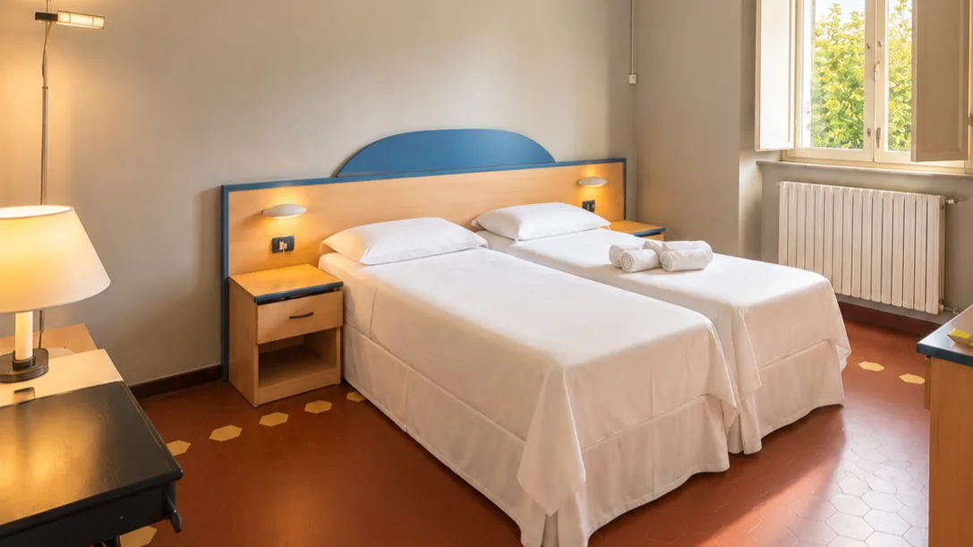 Twin bedroom with two single beds, blue and wood headboard, bedside tables, rolled towels and window overlooking greenery - Hotel Rex, BZAR hotels in Lucca