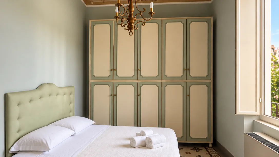 Bedroom with double bed, pale green tufted headboard, matching wardrobe, chandelier and rolled towels on white bedding - Hotel Rex, BZAR hotels in Lucca