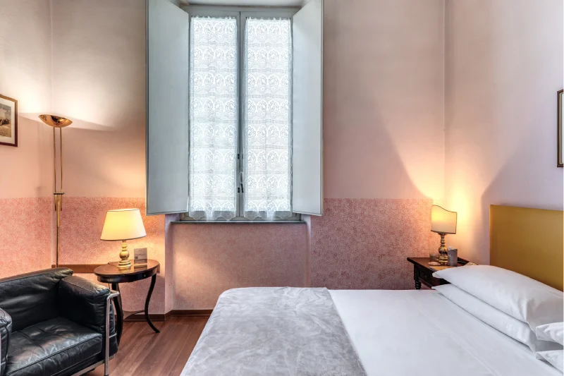 Double room with large bed, black armchair, desk, mirror and window with light curtains - Hotel Rex, BZAR hotels in Lucca