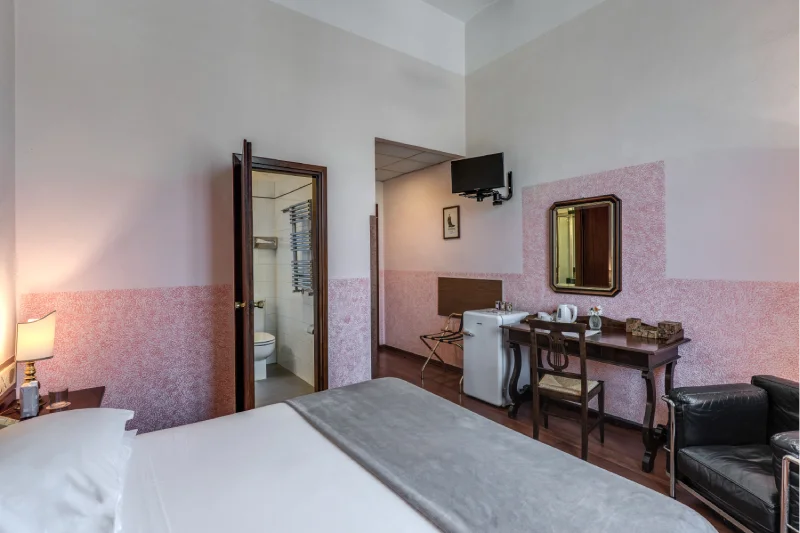 Double room with large bed, desk, mirror, TV, black armchair and visible private bathroom - Hotel Rex, BZAR hotels in Lucca