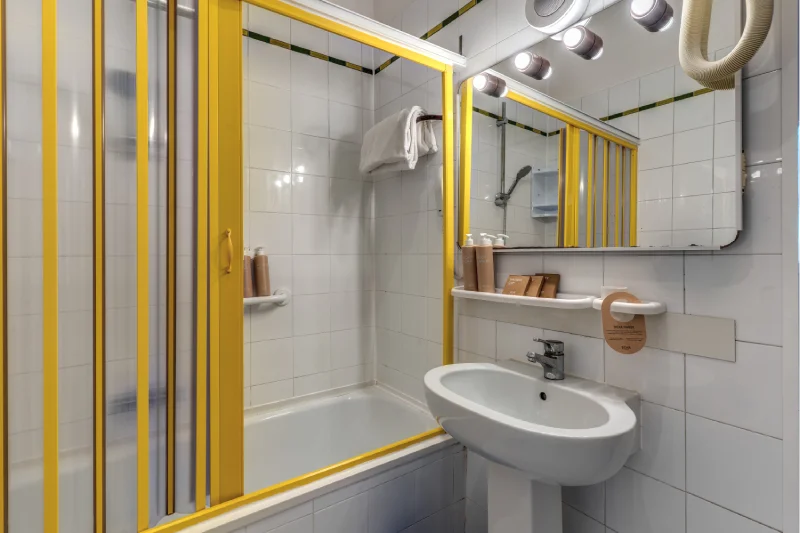 Family bathroom detail with yellow shower bath, white sink, mirror, lights and courtesy products - Hotel Rex, BZAR hotels in Lucca