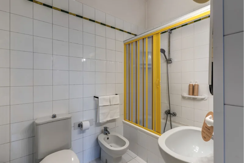 Family bathroom with shower bath, yellow panel, bidet, white sink and light tiles - Hotel Rex, BZAR hotels in Lucca