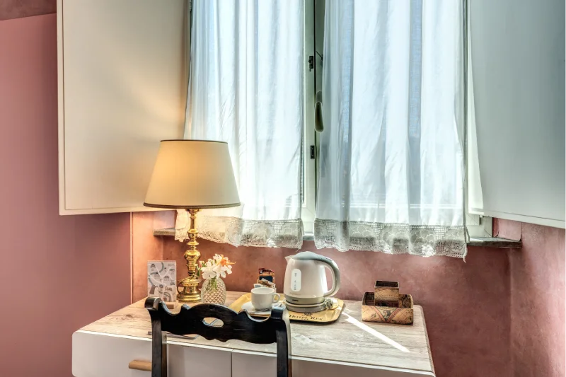 In-room desk detail with lamp, kettle, cup, amenities and bright window with curtains - Hotel Rex, BZAR hotels in Lucca