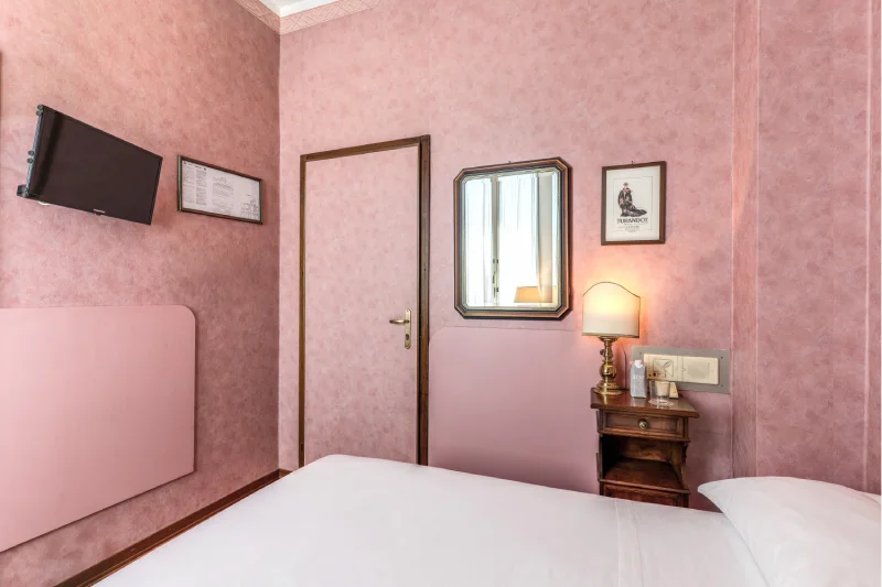 Single room with bed, wall-mounted TV, mirror, vintage bedside table, door and decorated pink walls - Hotel Rex, BZAR hotels in Lucca