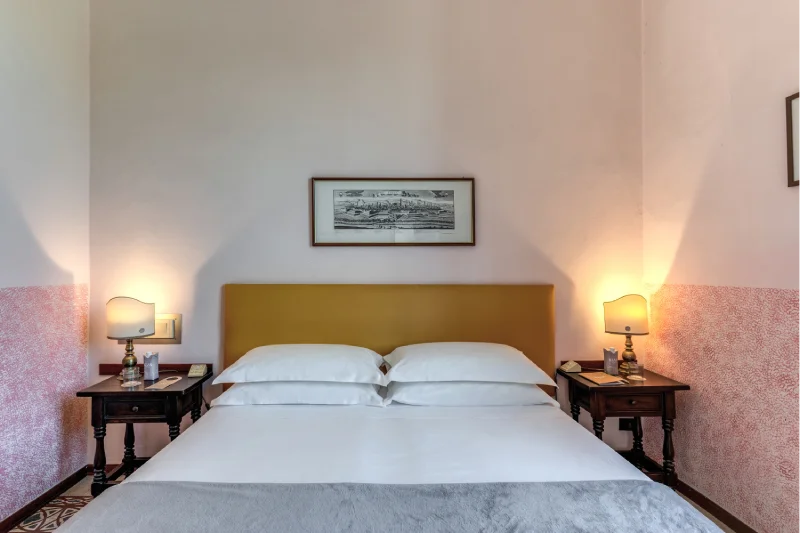 Suite with double bed, mustard headboard, vintage bedside tables, lit lamps and artwork above the bed - Hotel Rex, BZAR hotels in Lucca