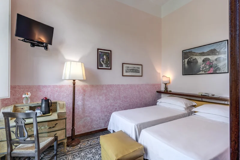 Suite with two single beds, wall-mounted TV, desk, kettle, floor lamp and pink walls - Hotel Rex, BZAR hotels in Lucca